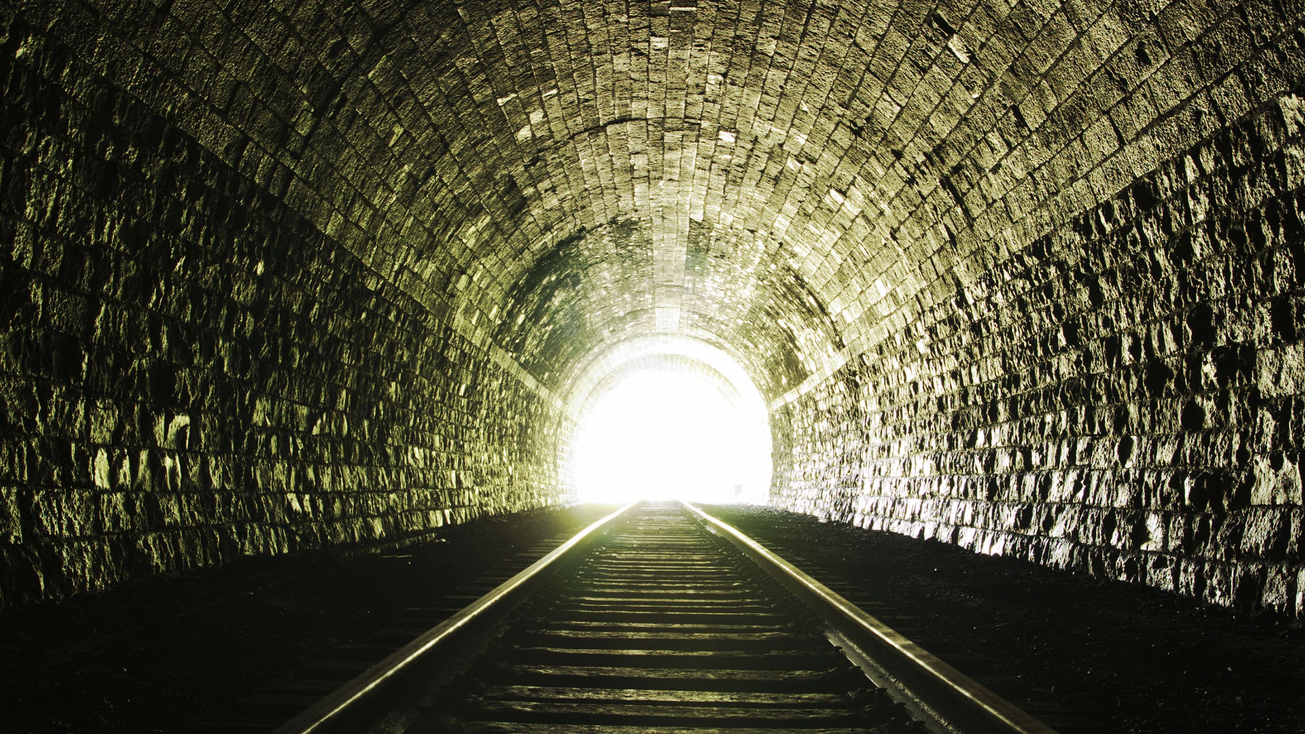 The image depicts the interior of a tunnel with rough stone walls, railway tracks leading toward a bright light at the far end, symbolizing hope and a journey from darkness to light.